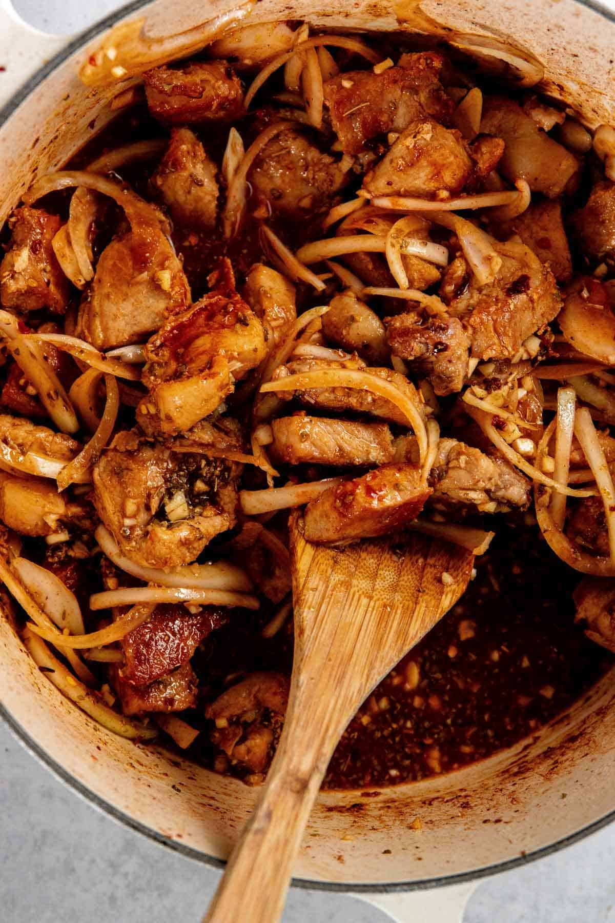 A pot of meat and veggies being stirred with a wooden spoon. The dish includes pork pieces, onions, and a dark, savory sauce.