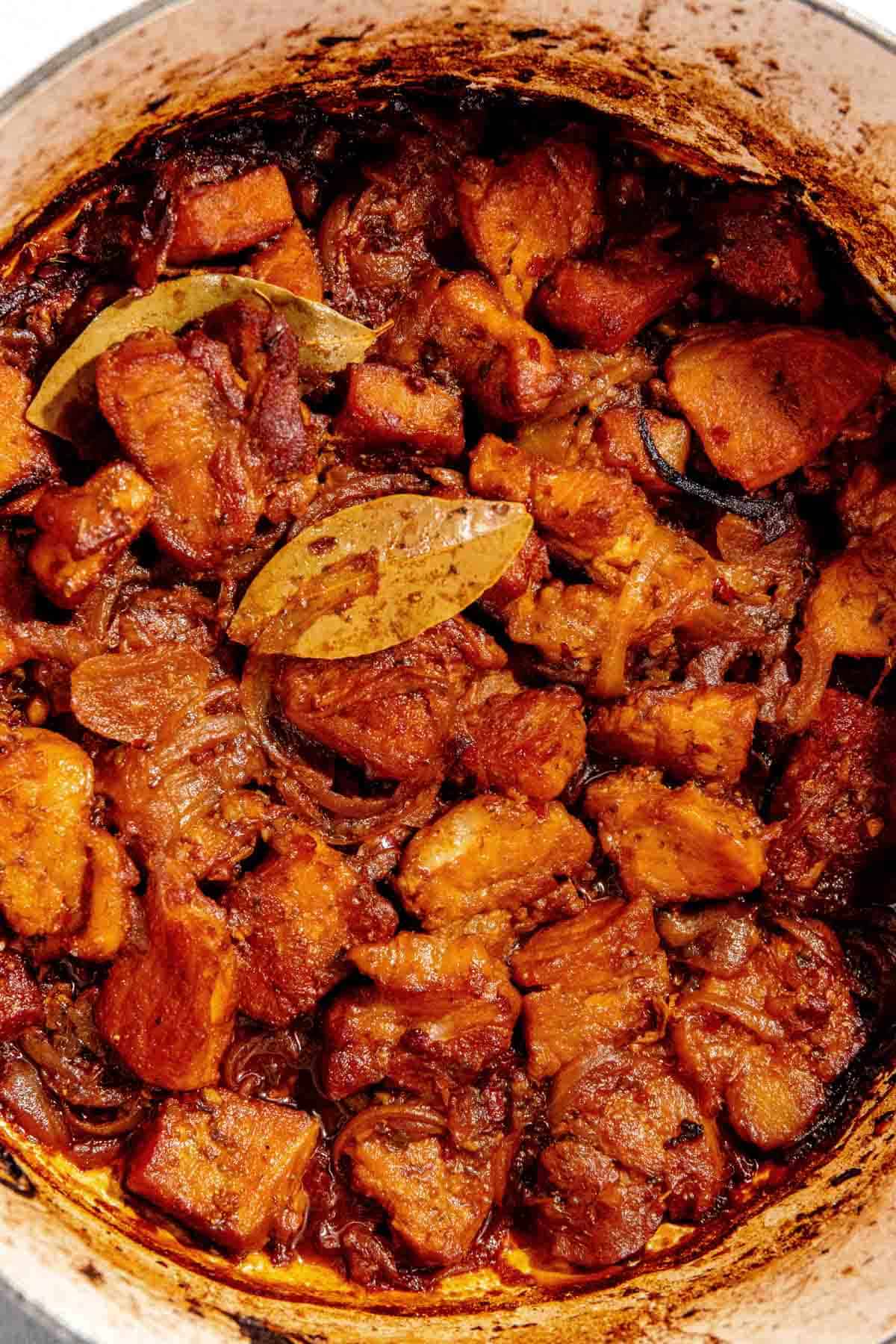 A close-up of a pot containing a braised carnitas made with chunks of meat, onions, and bay leaves, all cooked in a rich, dark sauce.