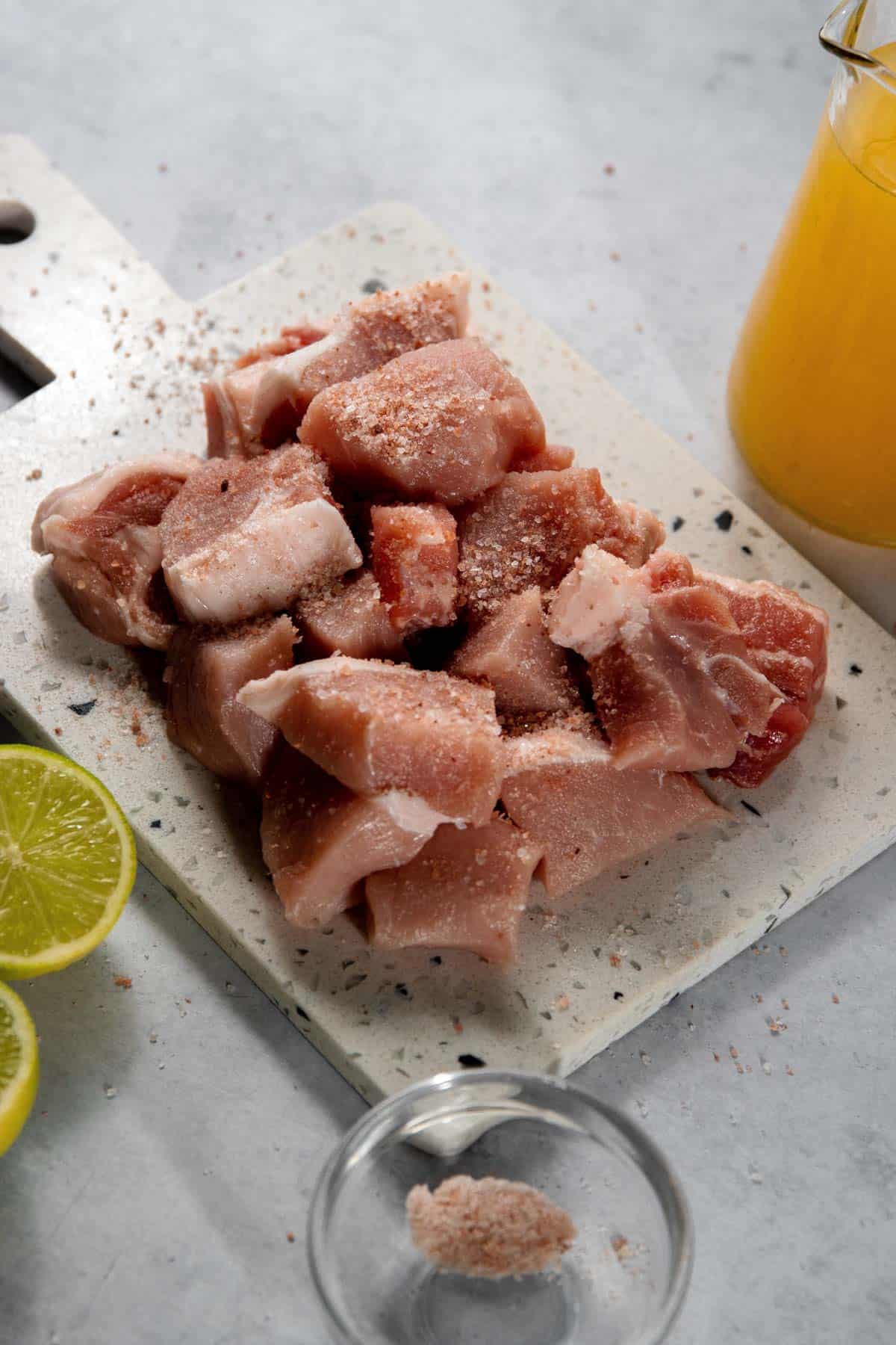 Chunks of raw pork seasoned with salt on a cutting board, accompanied by a lime, a small bowl of salt, and a jug of orange juice.