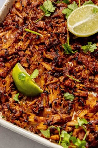 Close-up of a dish featuring shredded, browned jackfruit topped with cilantro and garnished with lime wedges.