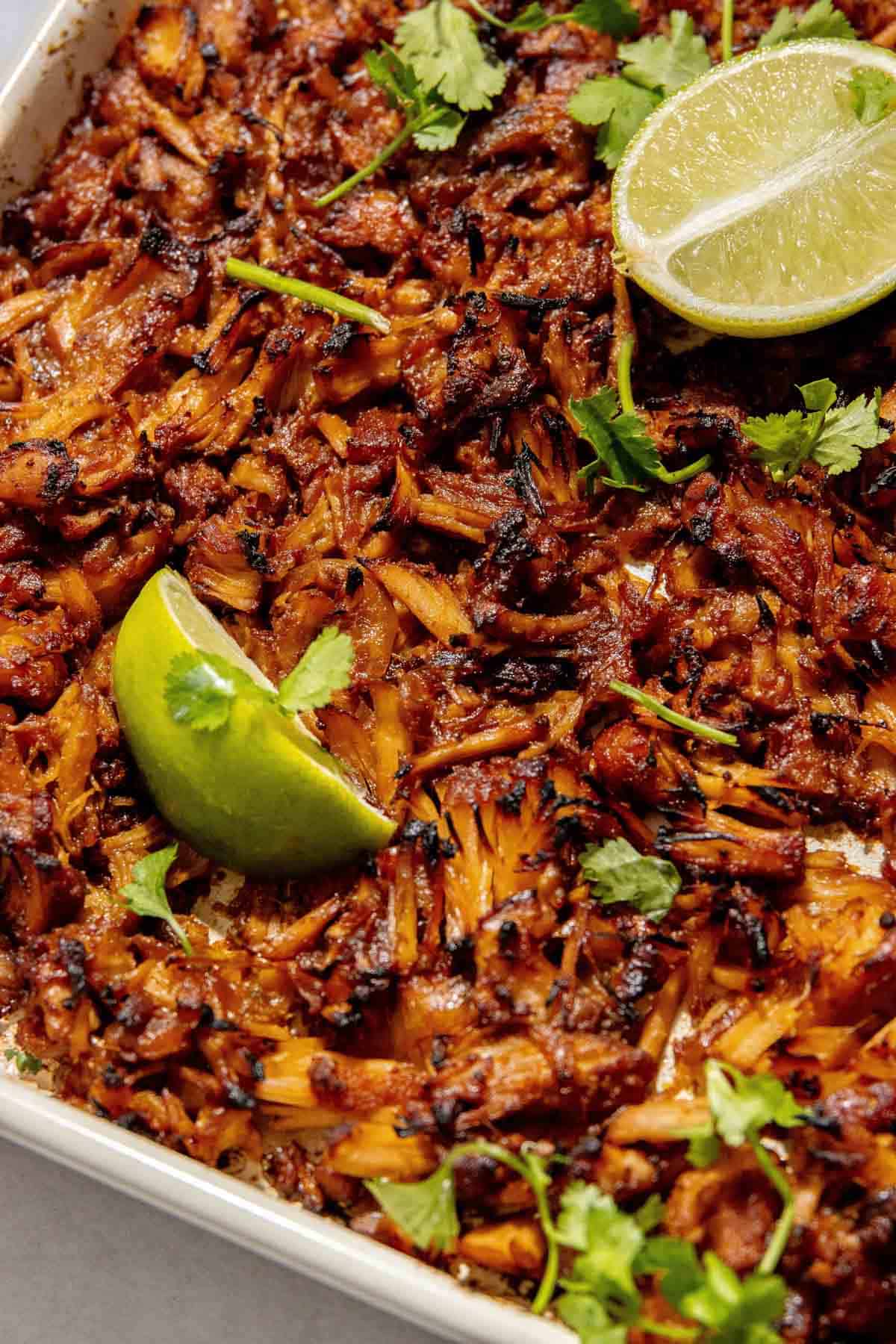 Close-up of a dish featuring shredded, browned carnitas topped with cilantro and garnished with lime wedges.