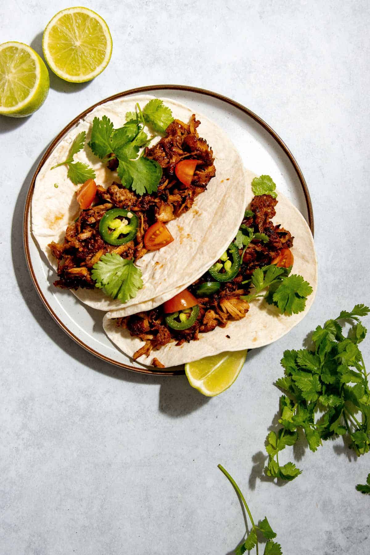 Two tacos on a plate filled with shredded pork canitas, jalapeños, tomato pieces, and cilantro. Garnished with lime halves and sprigs of cilantro on the side.