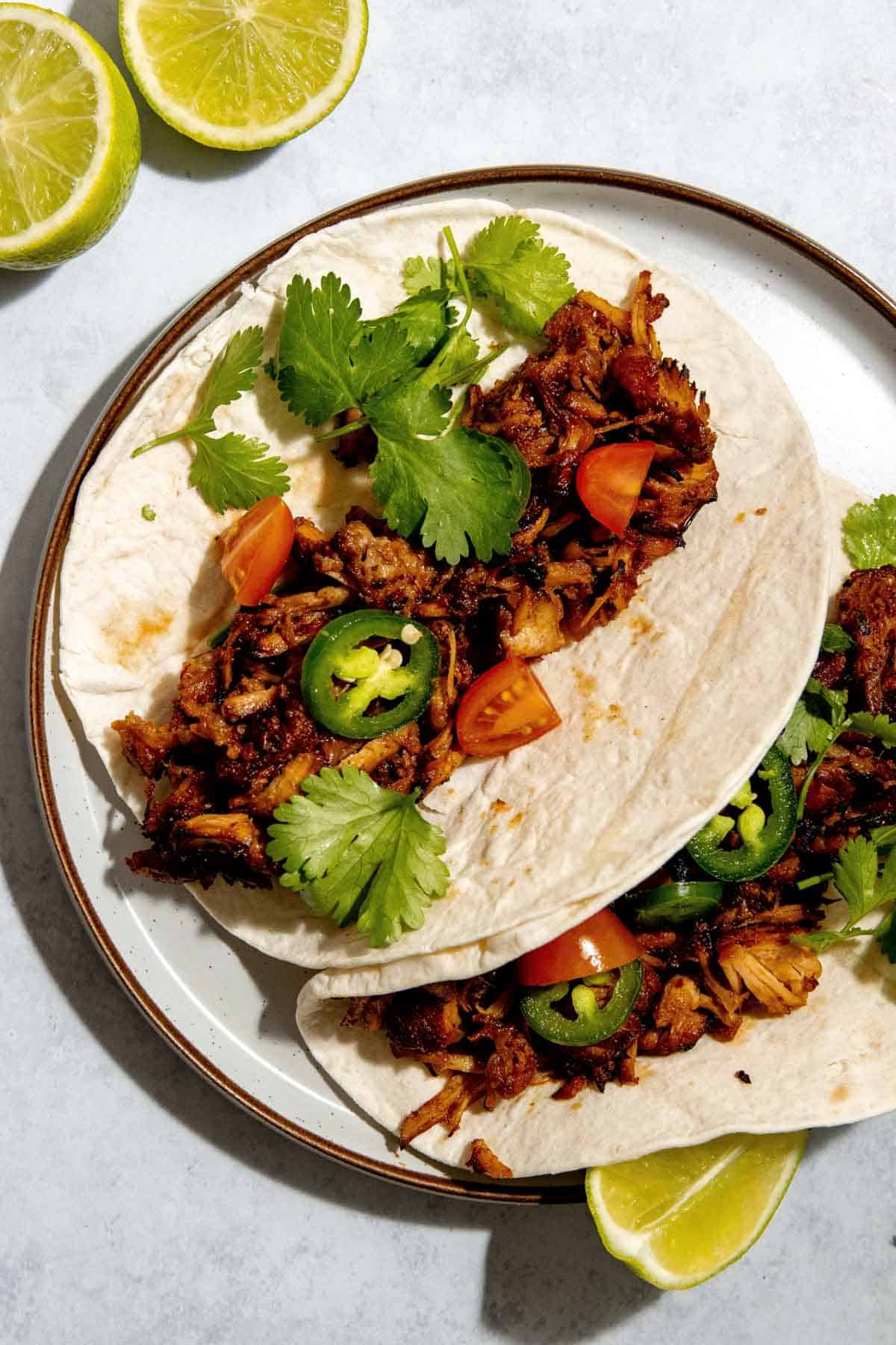 Two tacos on a plate filled with shredded pork carnitas, tomatoes, jalapeños, and cilantro. Sliced limes are visible on the side.