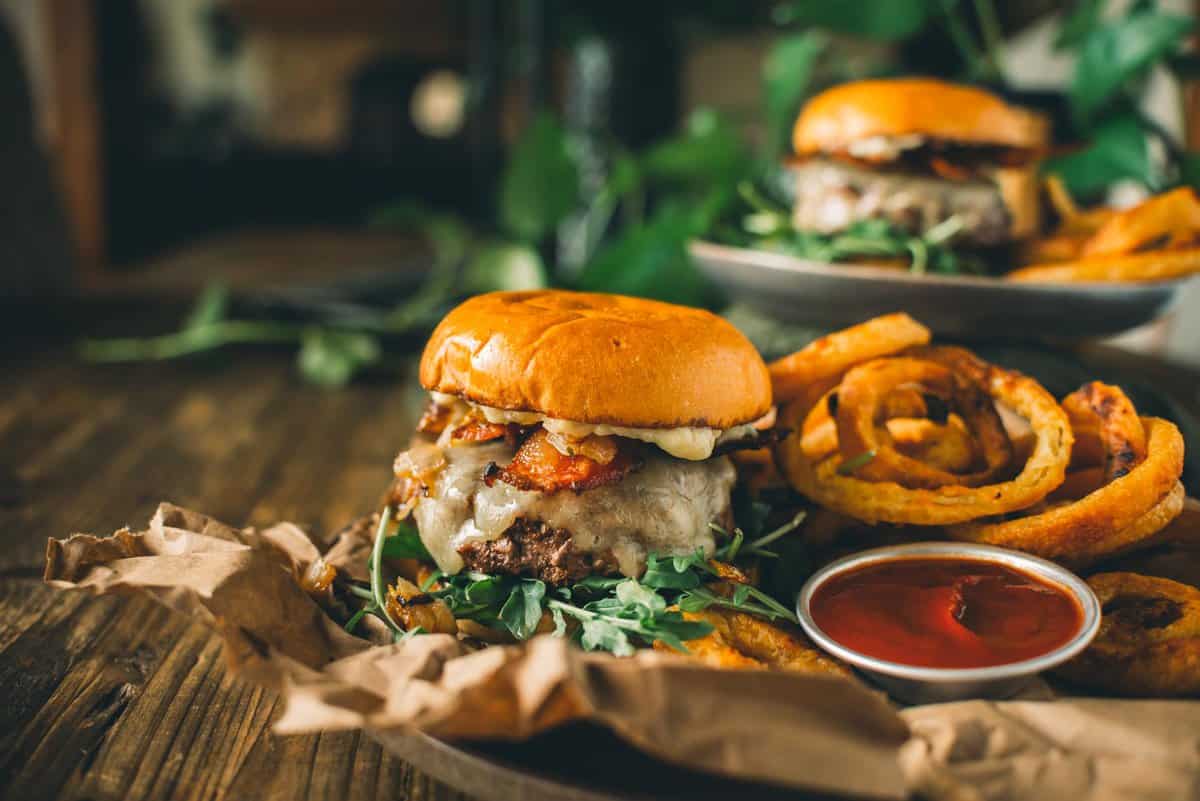 A cheeseburger with greens and caramelized onions sits on a rustic plate beside onion rings with a small cup of ketchup. Another similar dish is blurred in the background.