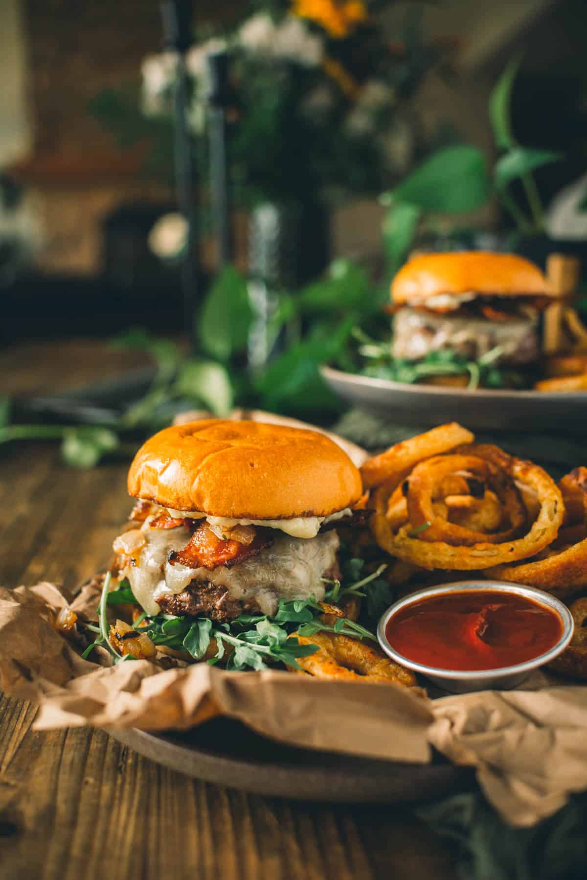 A bacon cheeseburger placed on a plate with curly fries and a side of ketchup on a wooden table. Another burger and decorative plants are visible in the blurred background.