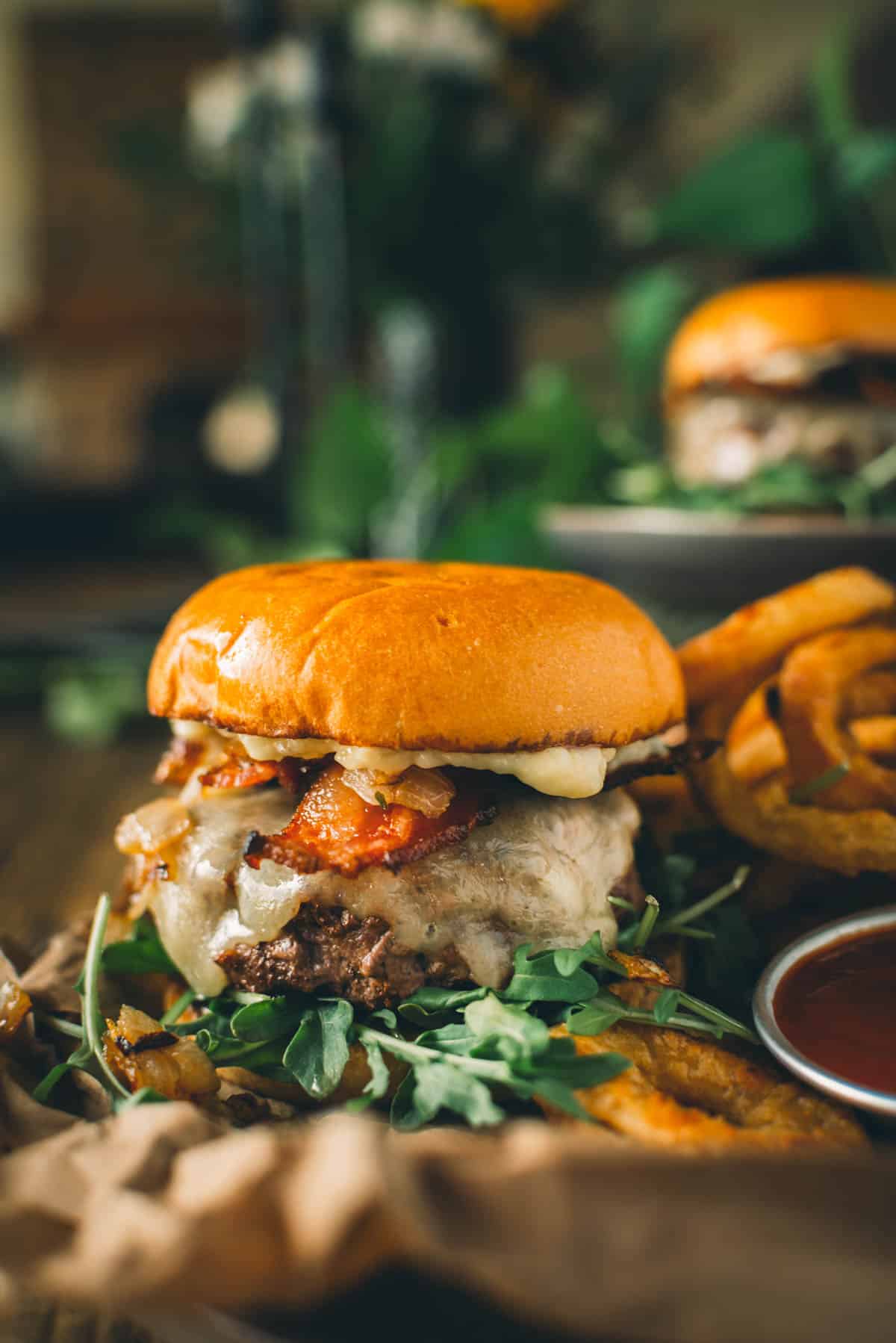 A garlic butter bacon cheeseburger with arugula, caramelized onions, and bacon on a bun, served with a side of curly fries and dipping sauce.