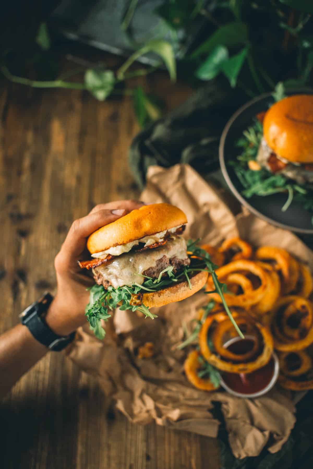 A hand holding a loaded bacon burger near a plate of crispy onion rings with a small container of dipping sauce, on a wooden surface with some greenery in the background.
