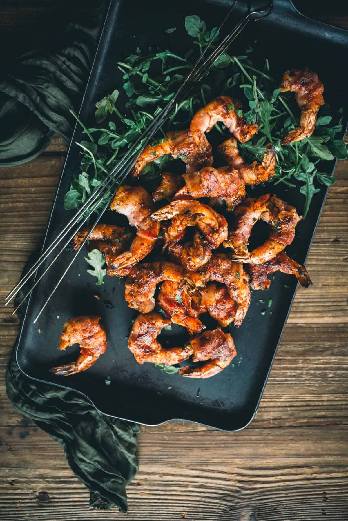 A tray of grilled shrimp wrapped in bacon placed on a bed of greens, with metal skewers on the side, on a wooden surface.
