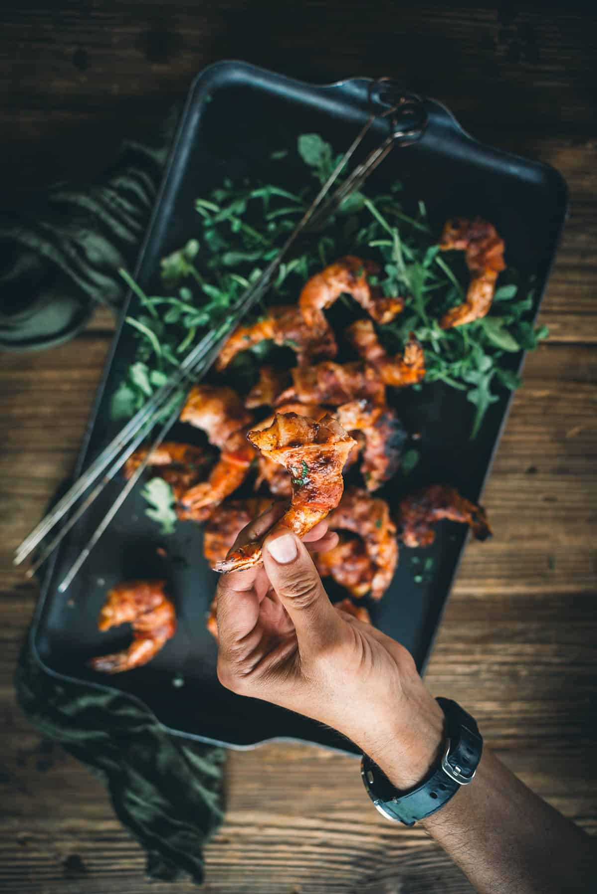 A person's hand holding a piece of grilled shrimp wrapped in bacon over a tray filled with more shrimp and greens on a wooden table.