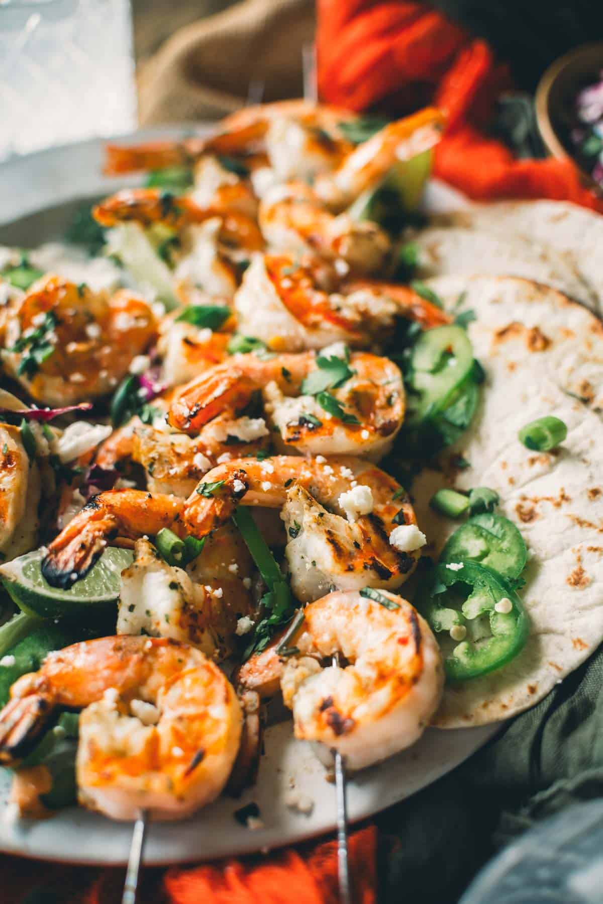 Grilled shrimp skewers topped with green onions and crumbled cheese, served with sliced jalapeños and tortillas on the side.