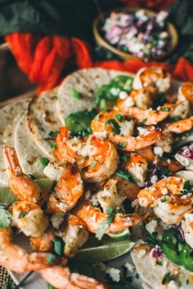Grilled shrimp on skewers, garnished with fresh herbs and served with tortillas and lime wedges, with a small bowl of cabbage slaw in the background.
