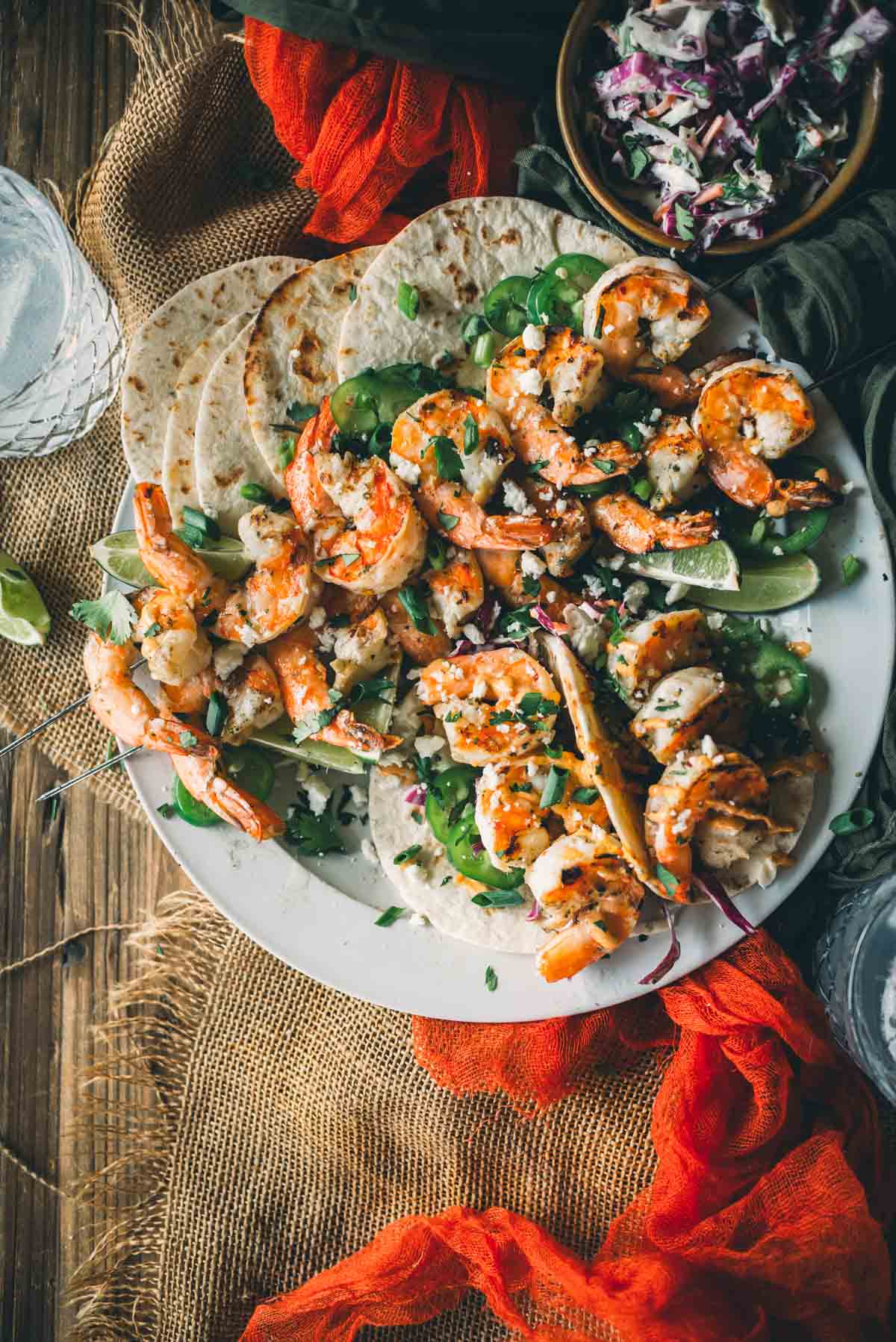 A plate of shrimp tacos with a side of coleslaw, garnished with herbs, displayed on a wooden table with a red cloth.