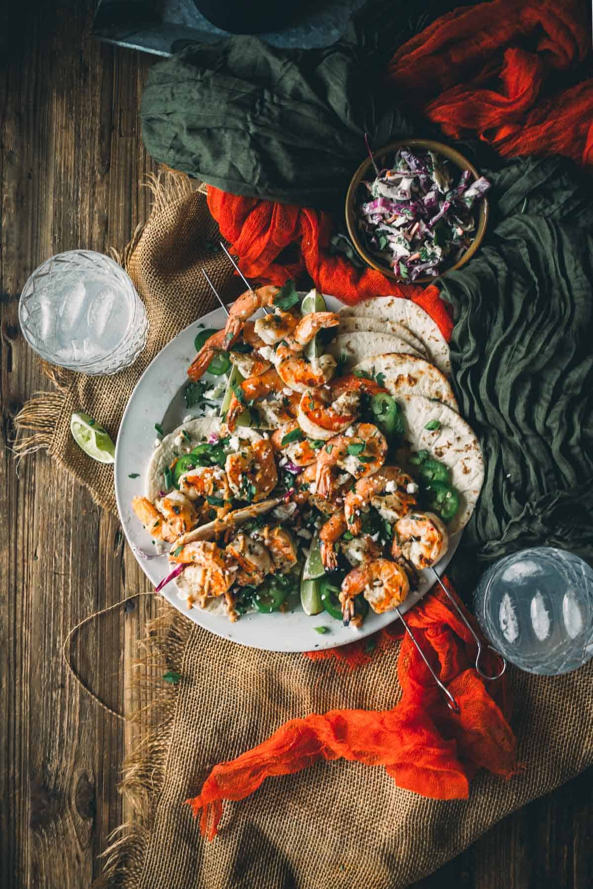 A plate of grilled shrimp, greens, and tortillas is placed on a wooden table with two margaritas and a small bowl of coleslaw. The setting includes burlap and red cloth accents.