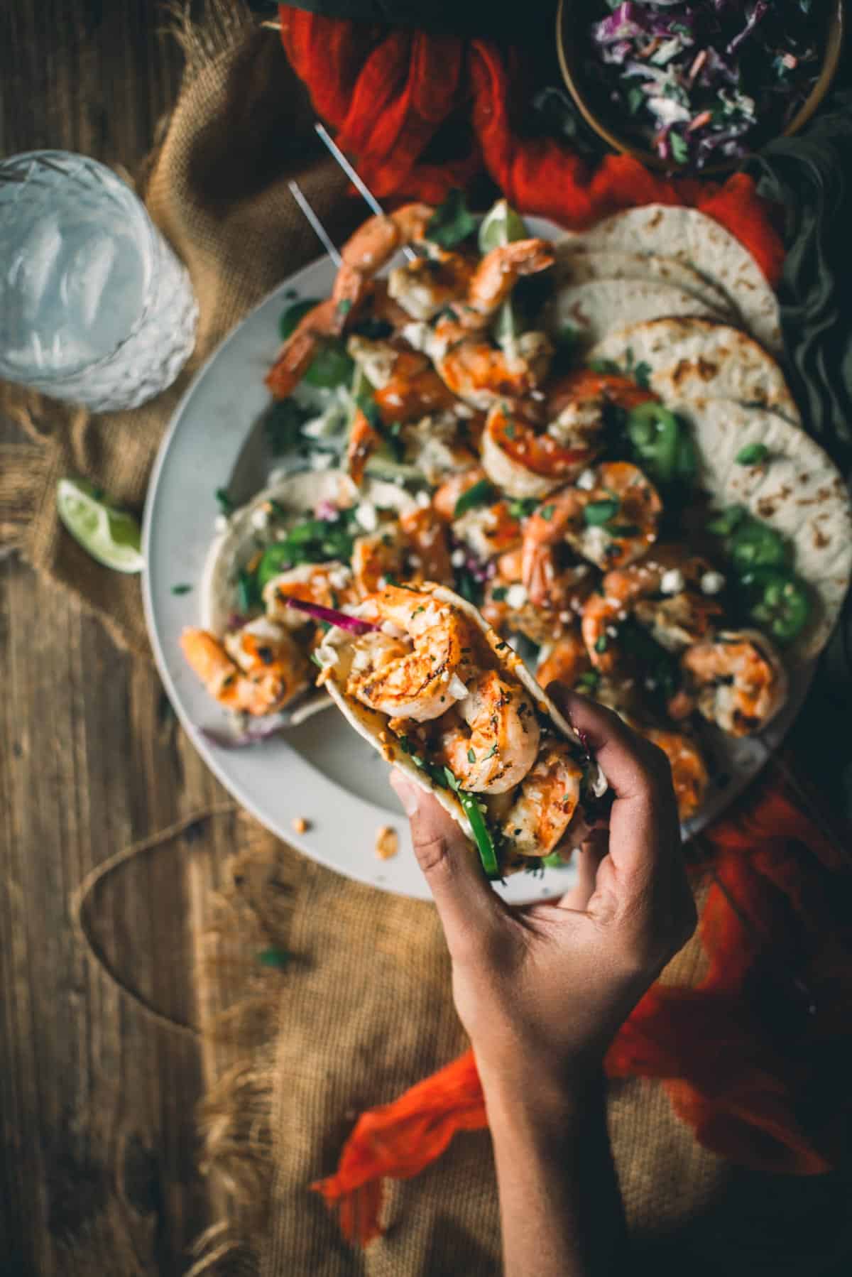 A hand holding a shrimp taco garnished with herbs and sauce, with more shrimp tacos and a drink on a rustic wooden table in the background.