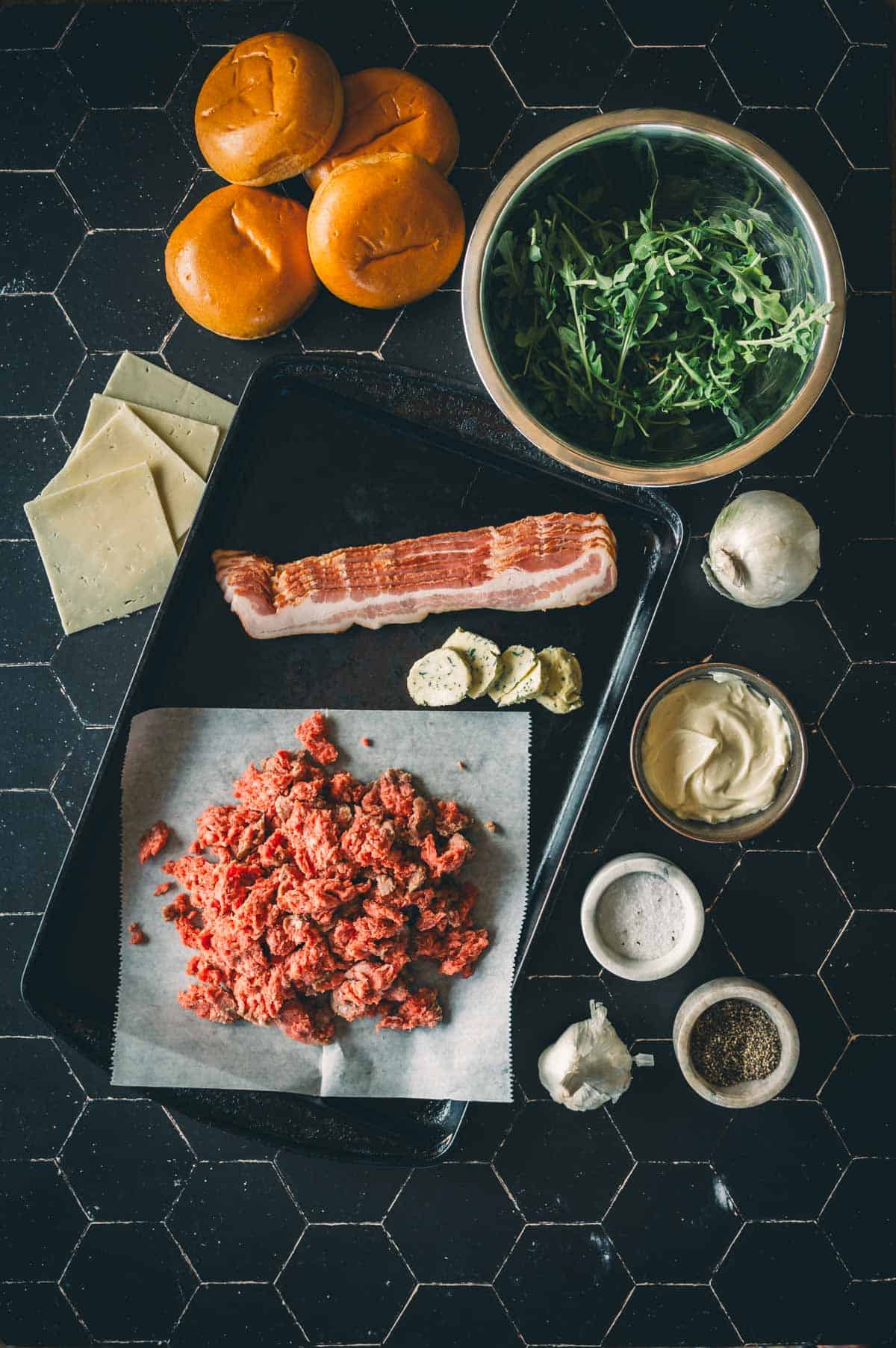 Ingredients for making burgers, including ground meat, bacon, cheese slices, brioche buns, arugula, garlic, salt, pepper, butter, and mayonnaise, arranged on a black surface.