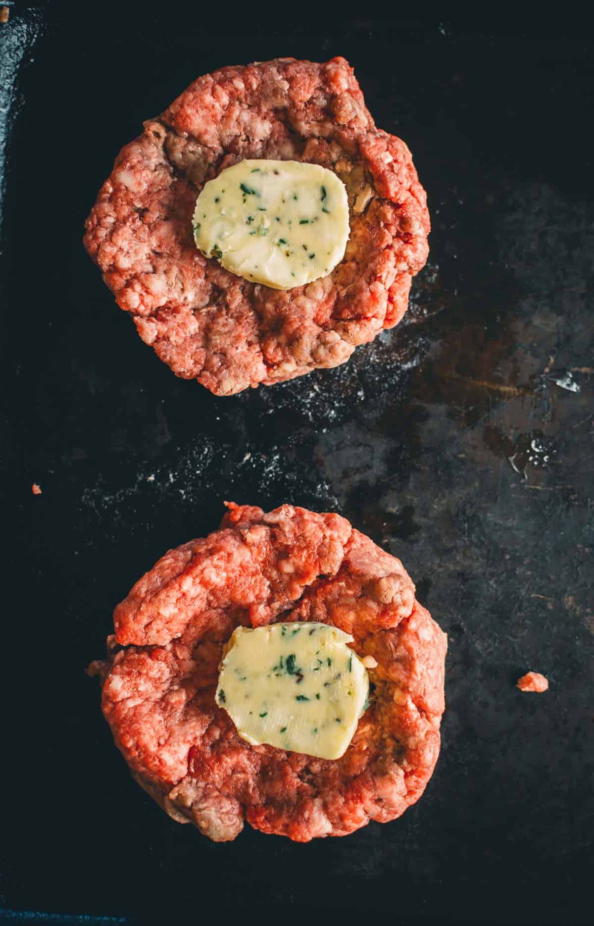 Two raw hamburger patties on a dark surface, each topped with a slice of herb butter.