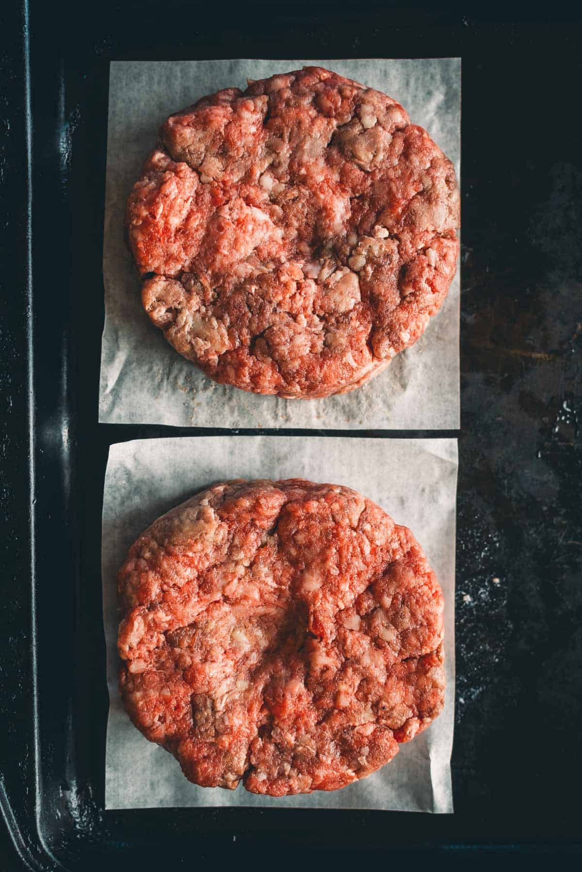 Two raw beef patties on parchment paper, placed on a dark baking tray with dimples in the center.