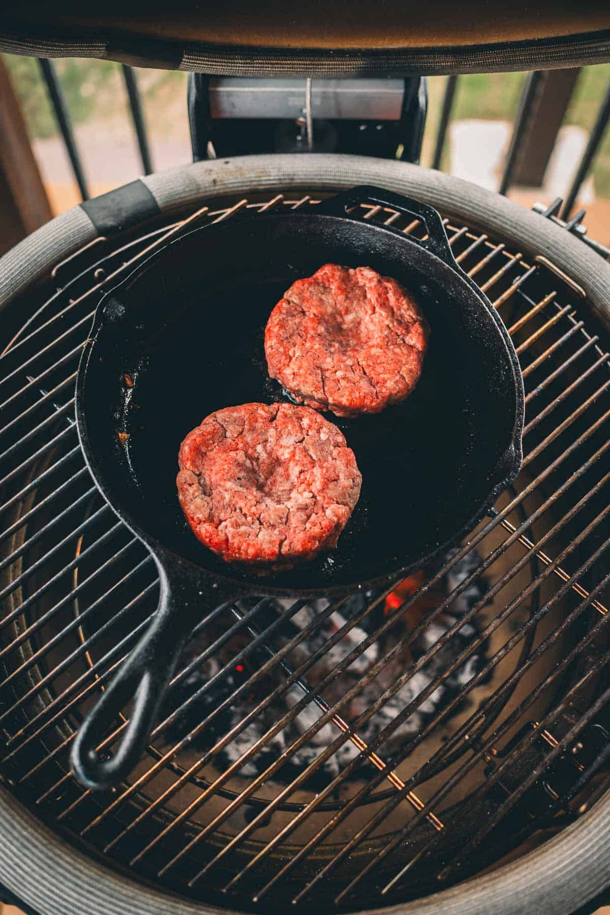Two raw hamburger patties are cooking in a cast iron skillet placed on a grill.