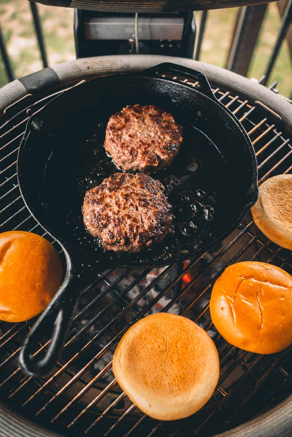 Two burger patties cooking in a cast iron skillet on a grill, with four hamburger buns toasting nearby on the grill grates.