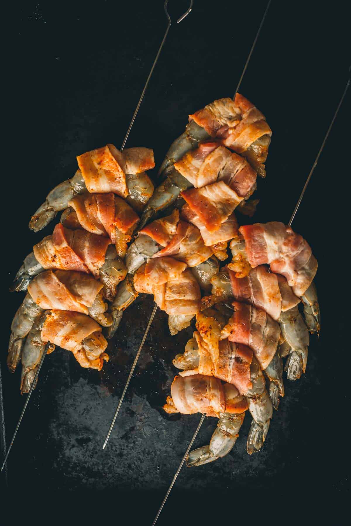 Skewers displaying raw shrimp wrapped with slices of bacon, arranged on a dark surface.