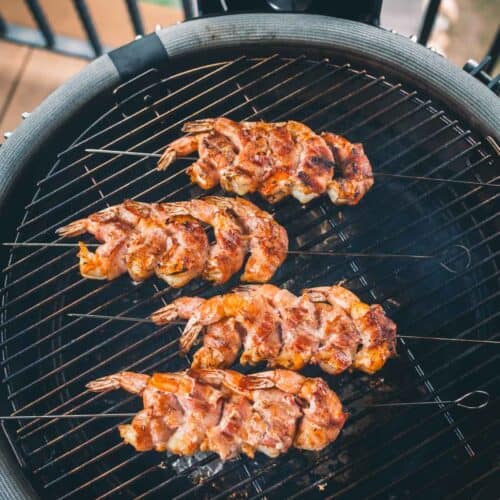 Four skewers of bacon wrapped shrimp are grilling on a barbecue grill.