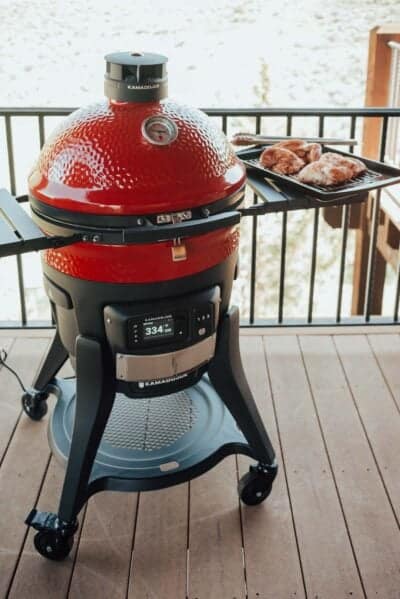A red Kamado Joe grill is on a wooden deck, with a tray of seasoned chicken pieces placed on its side shelf.