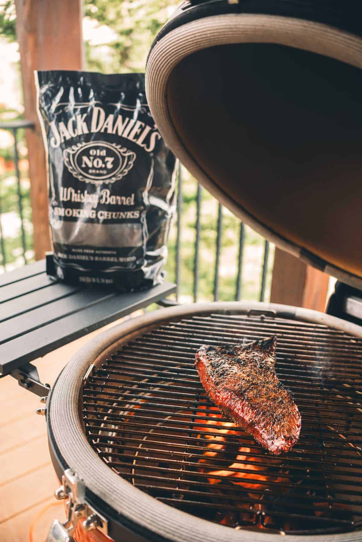 A seasoned steak cooks on a grill with glowing coals beneath. A bag of Jack Daniel's Whiskey Barrel Smoking Chunks is placed next to the grill.
