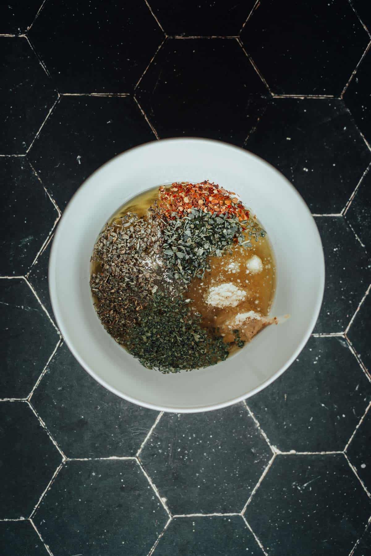 A white bowl on a black hexagon-tiled surface contains various herbs and seasonings pre-mixed together.
