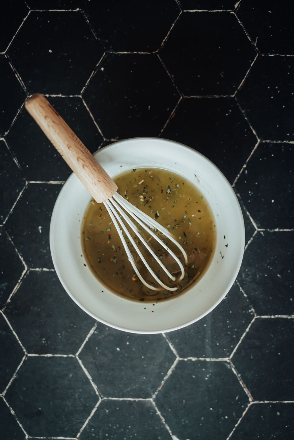 A bowl of homemade italian dressing with herbs, being mixed with a whisk, sits on a black surface.