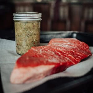 A raw steak sits on parchment paper with a jar of marinade beside it, both placed on a dark tray in a kitchen.