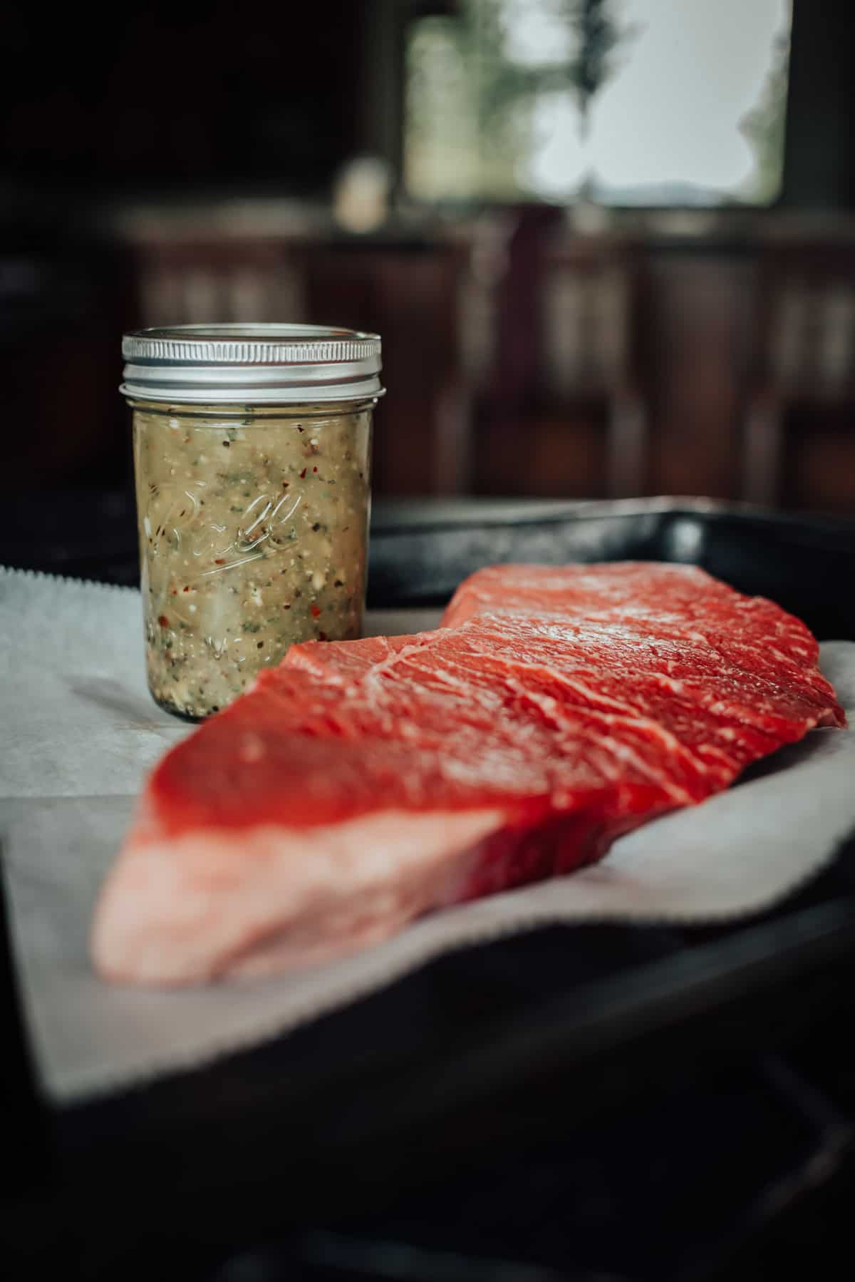 A raw steak sits on parchment paper with a jar of marinade beside it, both placed on a dark tray in a kitchen.