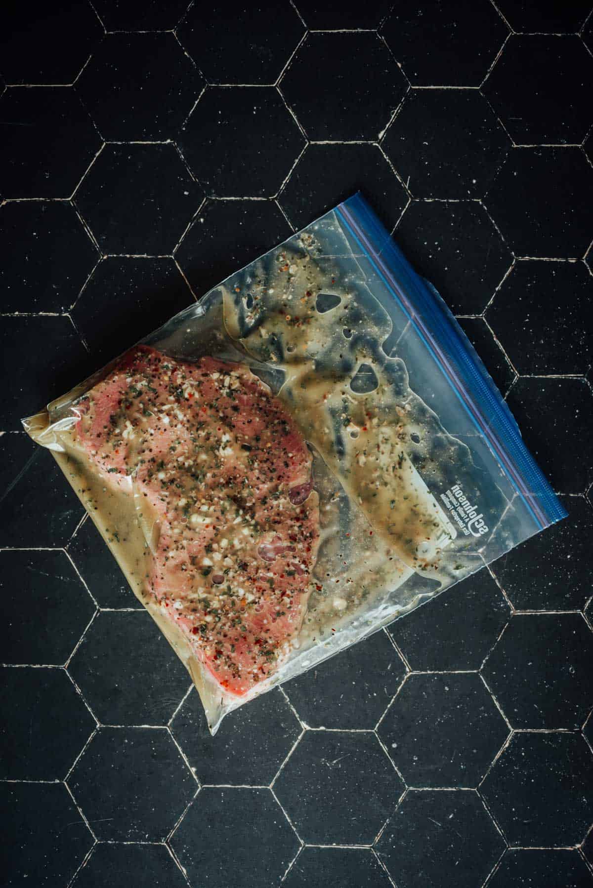 A londlon broil marinating in a resealable plastic bag lies on a dark hexagonal tile surface.