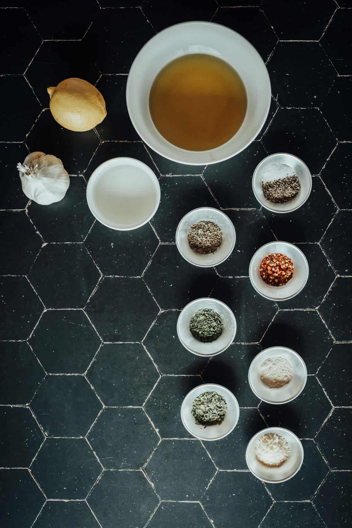 Top view of a variety of ingredients in bowls on a black hexagonal tile surface, including spices, a lemon, garlic, and a bowl of liquid.
