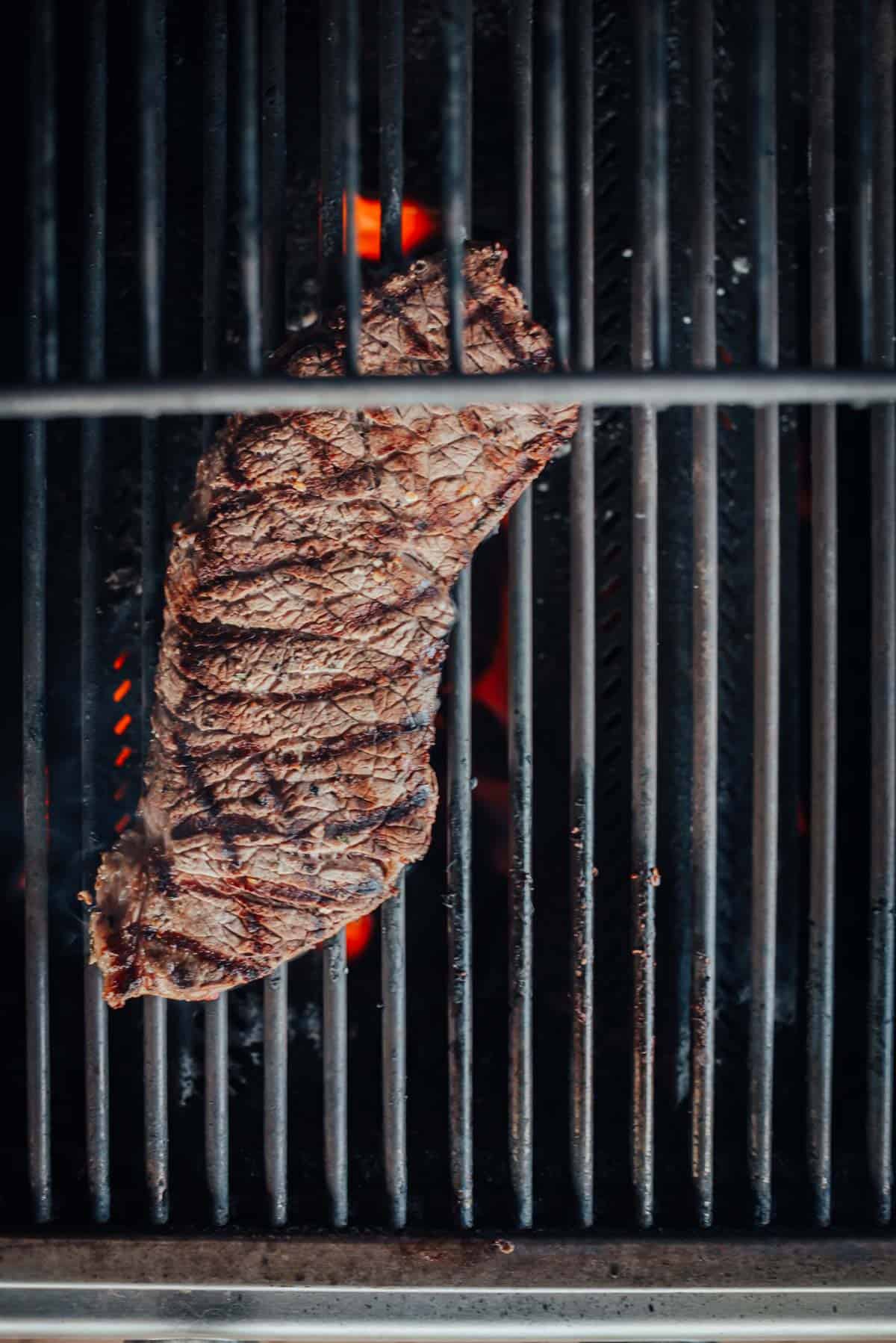 A london broil is being grilled on a barbecue, showing grill marks and flames beneath the grates.