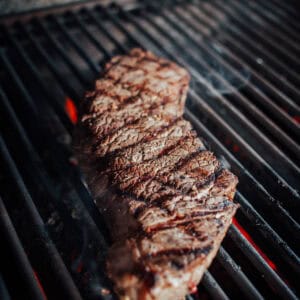 A london broil being grilled on an open flame grill, with visible char marks and red-hot coals underneath.