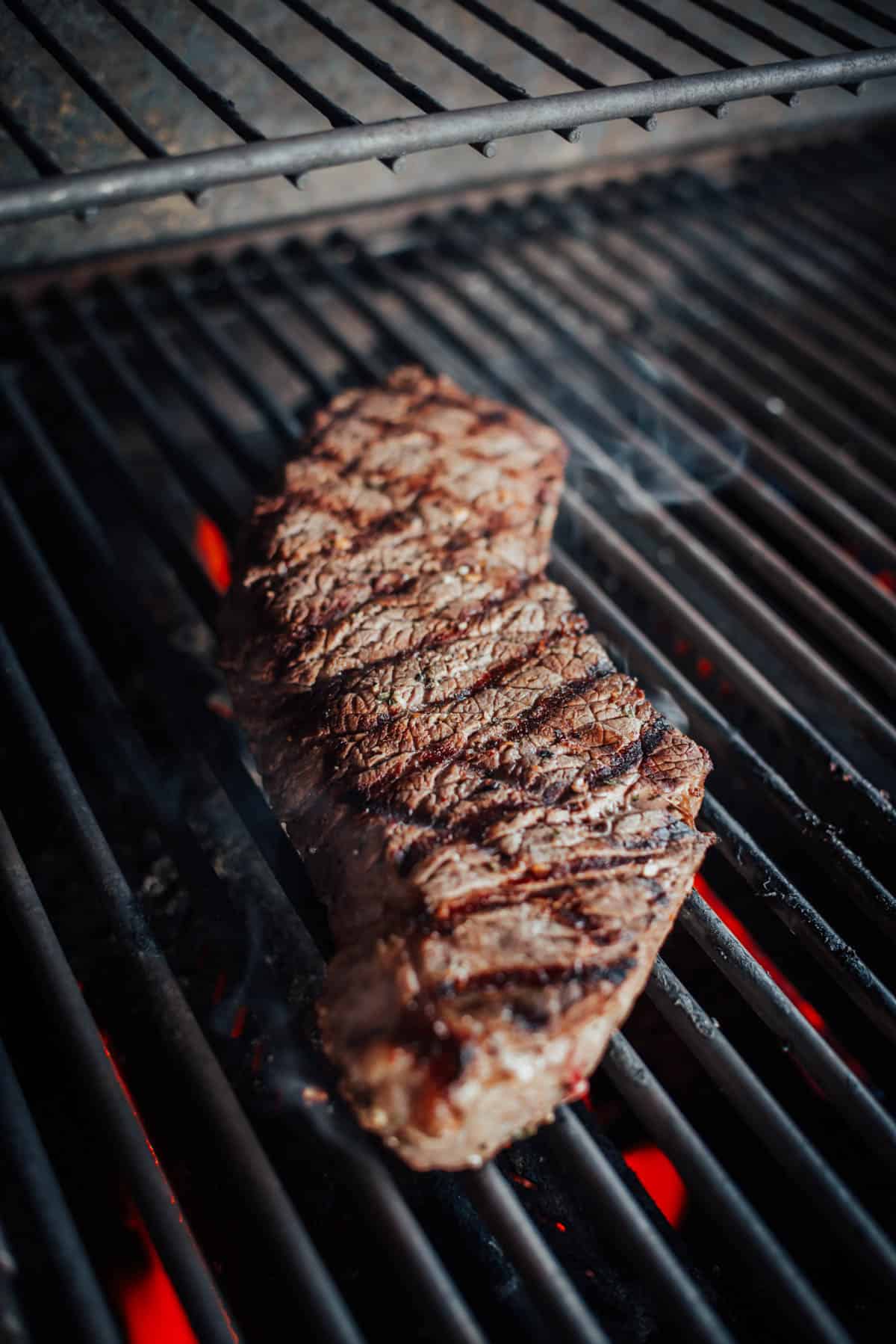 A london broil being grilled on an open flame grill, with visible char marks and red-hot coals underneath.