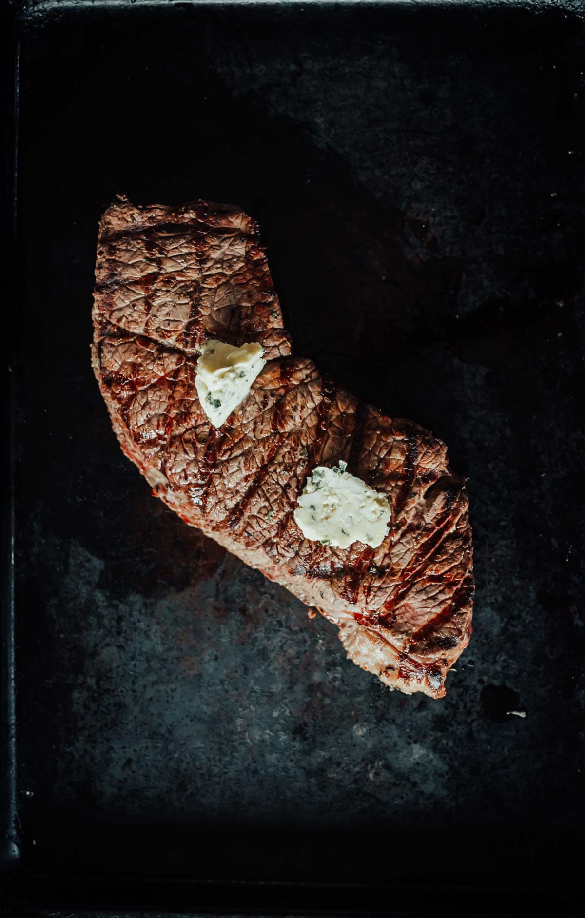 A grilled steak resting with two small pats of melting butter on top, placed on a dark surface.