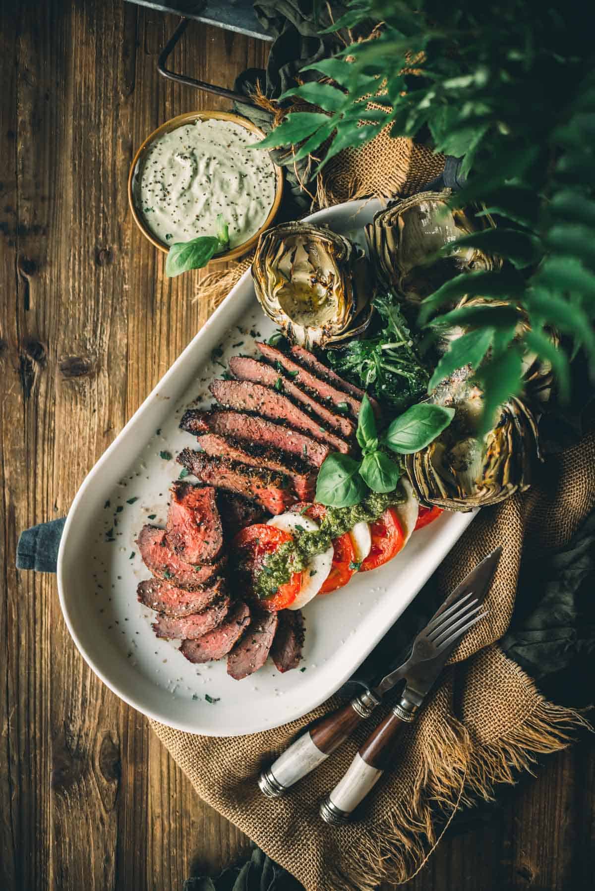 A platter with sliced tri-tip, fresh tomatoes, mozzarella, basil leaves, and grilled artichokes. Two forks on the side. A bowl filled with a green dip in the background.