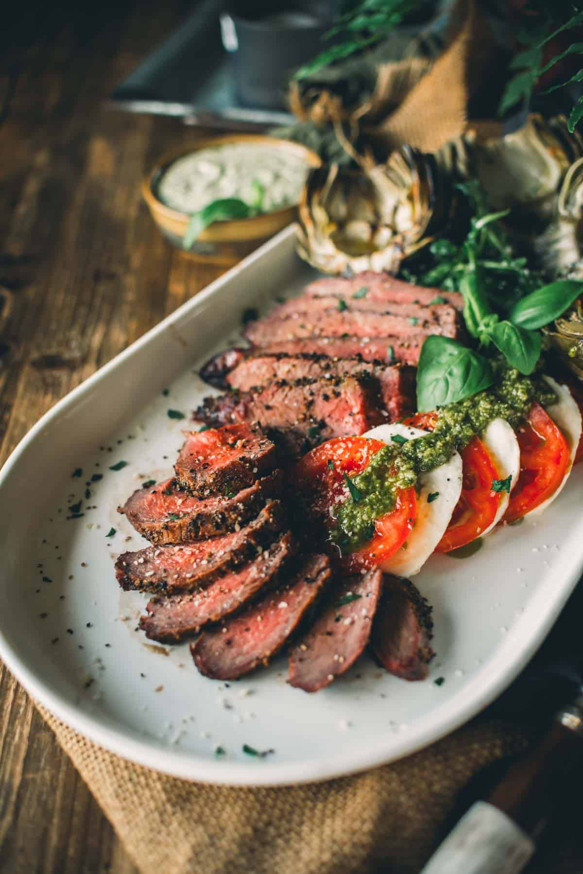 A white plate with sliced smoked tri-tip, tomato slices, mozzarella, basil, drizzled with green sauce, and a side of dipping sauce in the background.