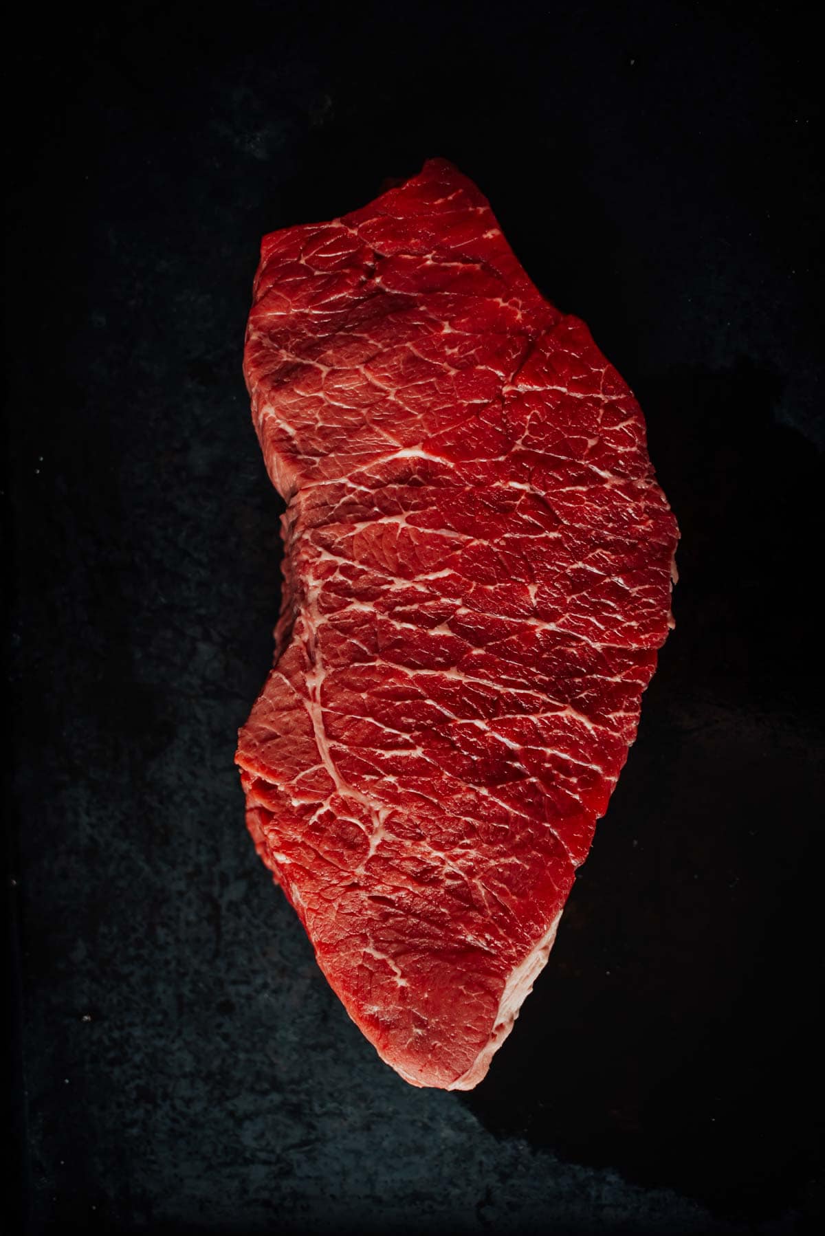 A raw, london broil steak against a dark background. The meat has a deep red color with visible marbling.