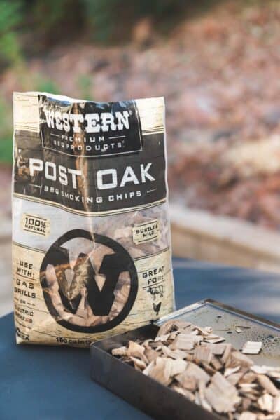 Bag of Western Premium Products Post Oak BBQ Smoking Chips placed on a surface next to a metal container filled with wood chips.