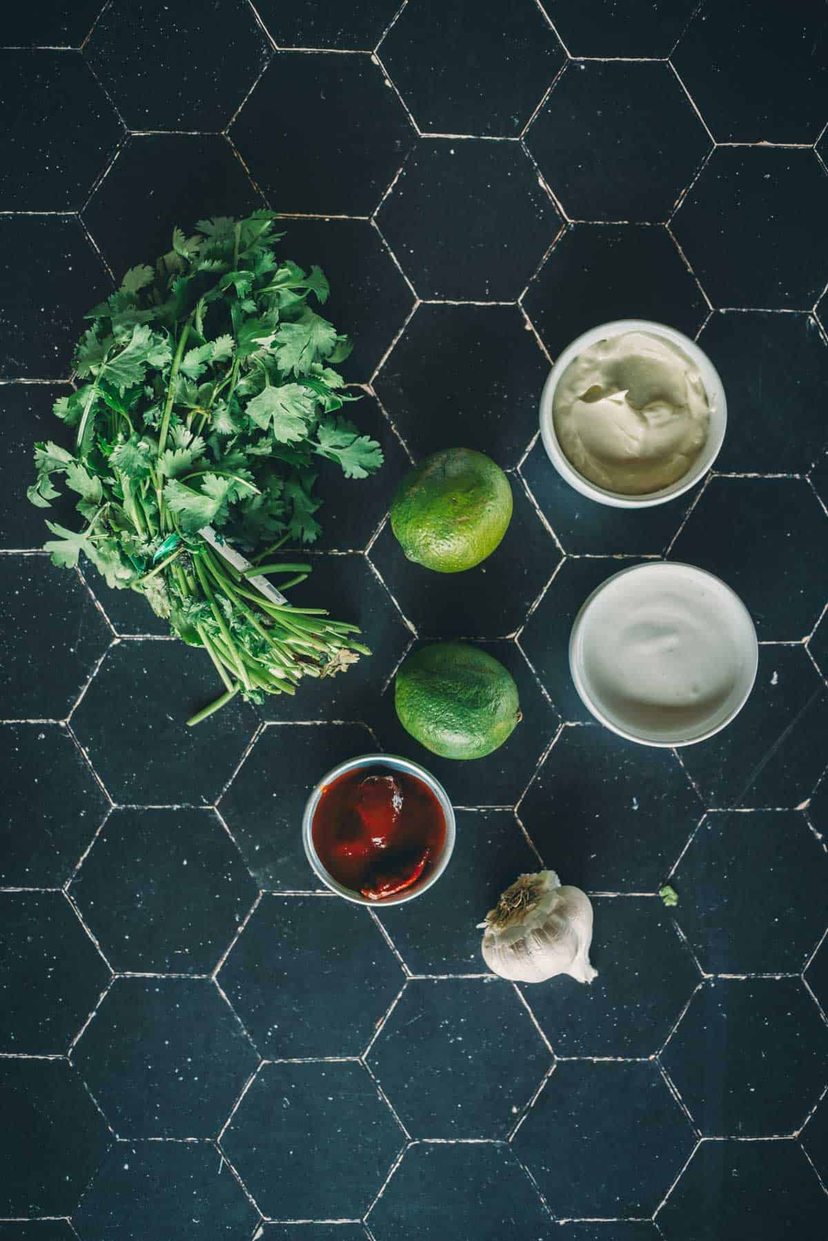 Cilantro, two limes, garlic, chipotle chiles in adobe, mayo, and crema are arranged on a black countertop.