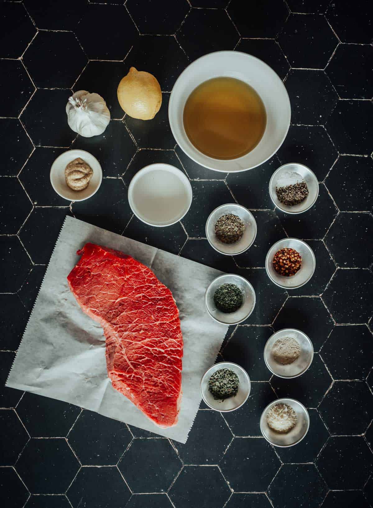 Raw london broil on parchment paper surrounded by bowls of spices, a garlic bulb, a lemon, and a bowl of oil, all placed on a black surface.