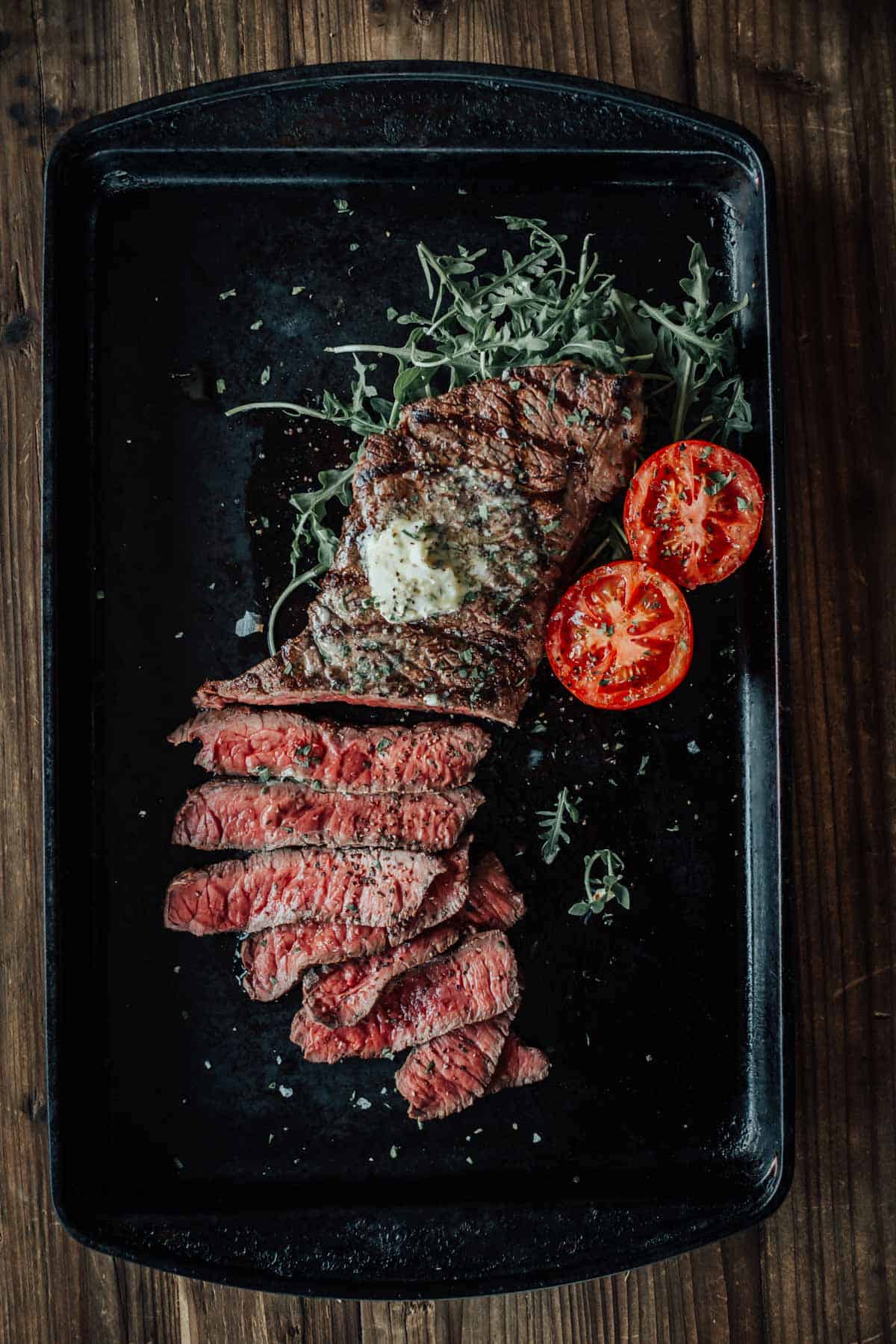A grilled london broil, sliced and topped with a dollop of butter, is served on a black tray with halved grilled tomatoes and a bed of arugula.