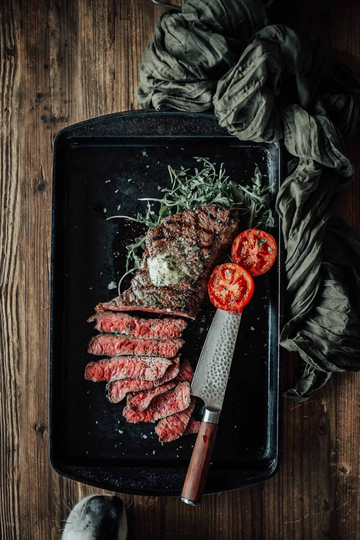 A grilled london broil with half sliced into pieces, garnished with a pat of butter, vegetables, and two grilled tomato halves, served on a black tray with a knife and a dogs nose sneaking in near the edge for a sniff.
