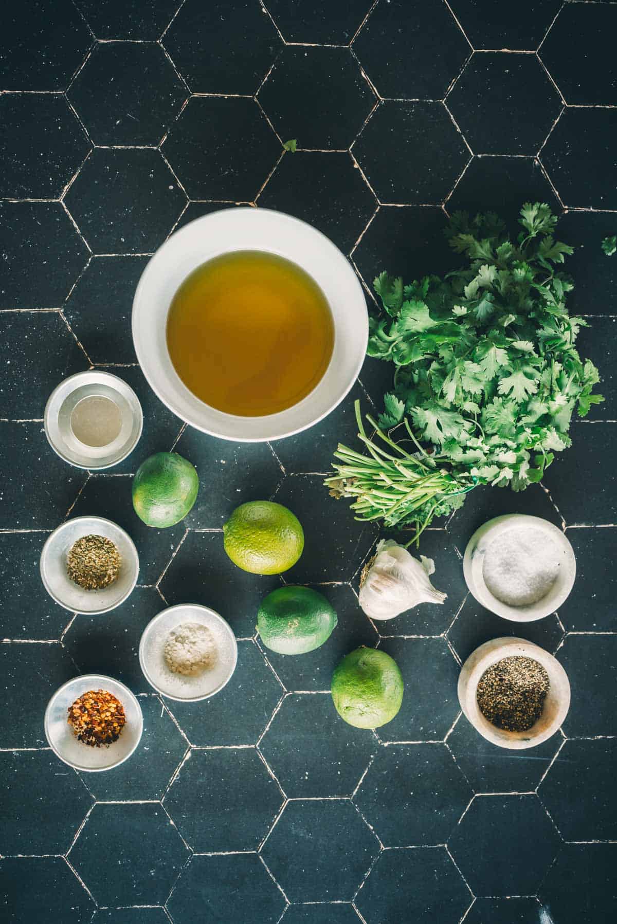 Overhead view of a table with various cooking ingredients, including a bowl of oil, bunch of cilantro, limes, garlic, and small bowls containing spices and seasonings.