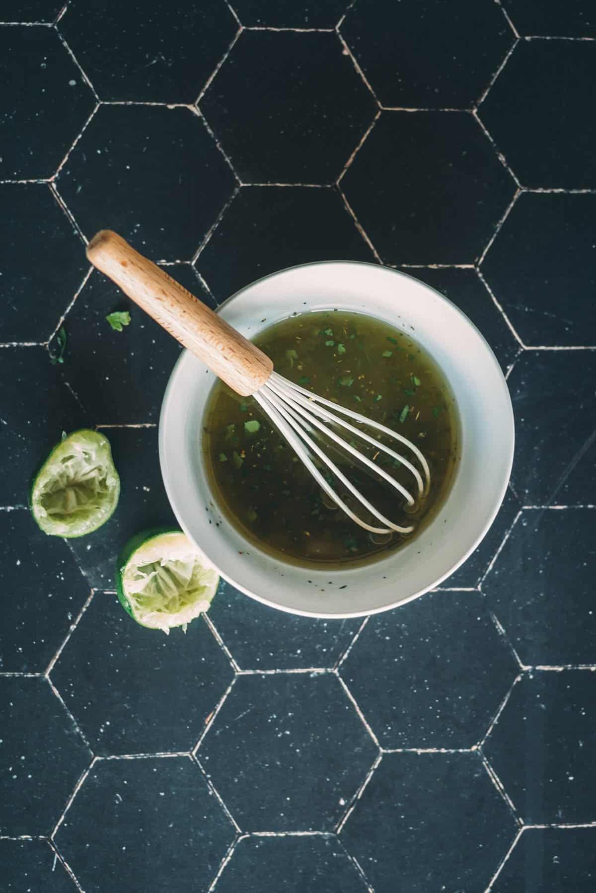 A bowl filled with a marinade, a whisk placed inside it, and two squeezed lime halves on a black hexagon-tiled surface.