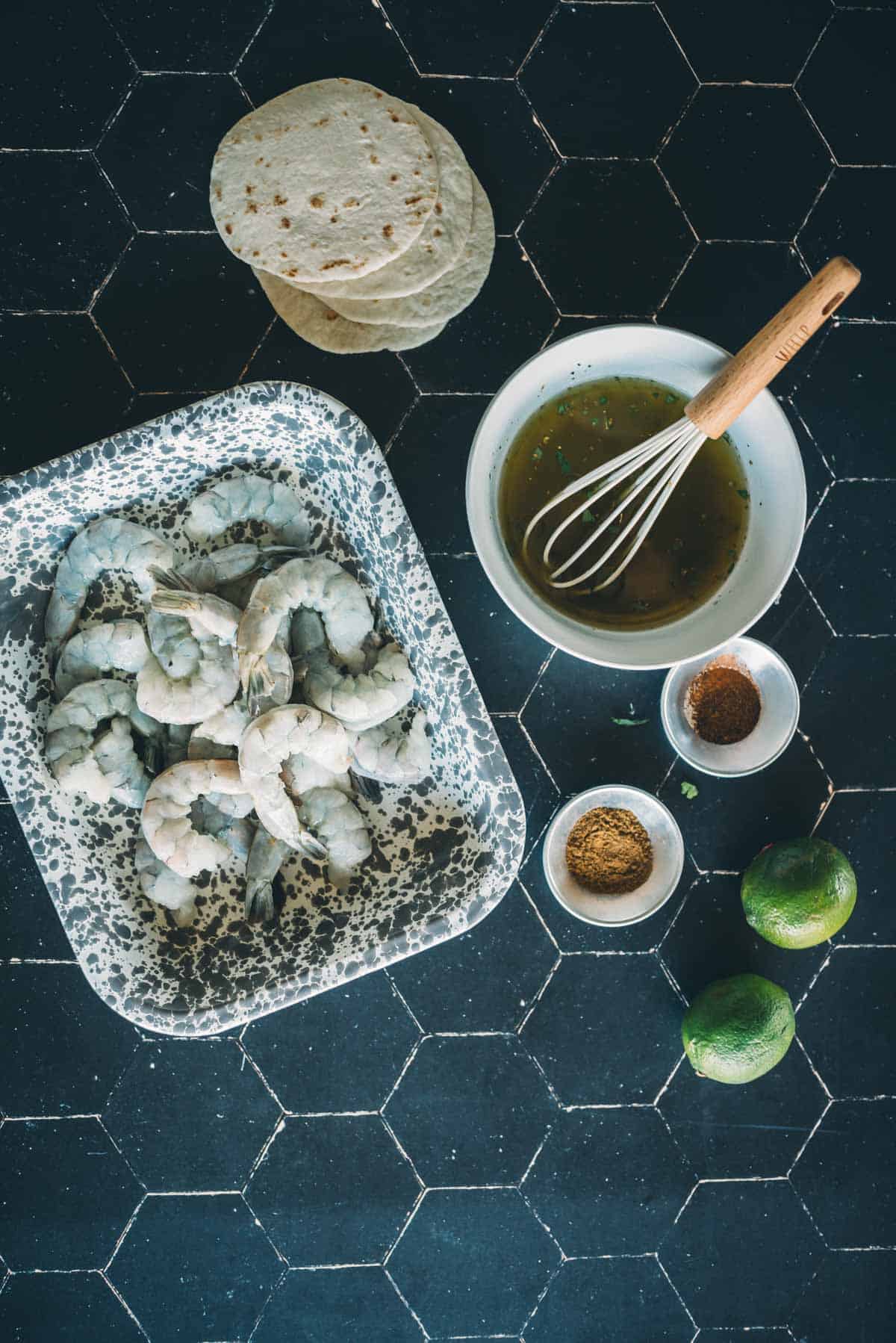 A kitchen counter with raw shrimp, spice bowls, lime, a whisk in a bowl of sauce, and tortillas.