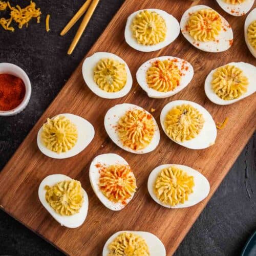 A wooden board with several deviled eggs topped with a yellow filling, some sprinkled with paprika, alongside a small bowl of paprika and two gold spoons—perfect for adding a festive touch to your 4th of July grilling spread.