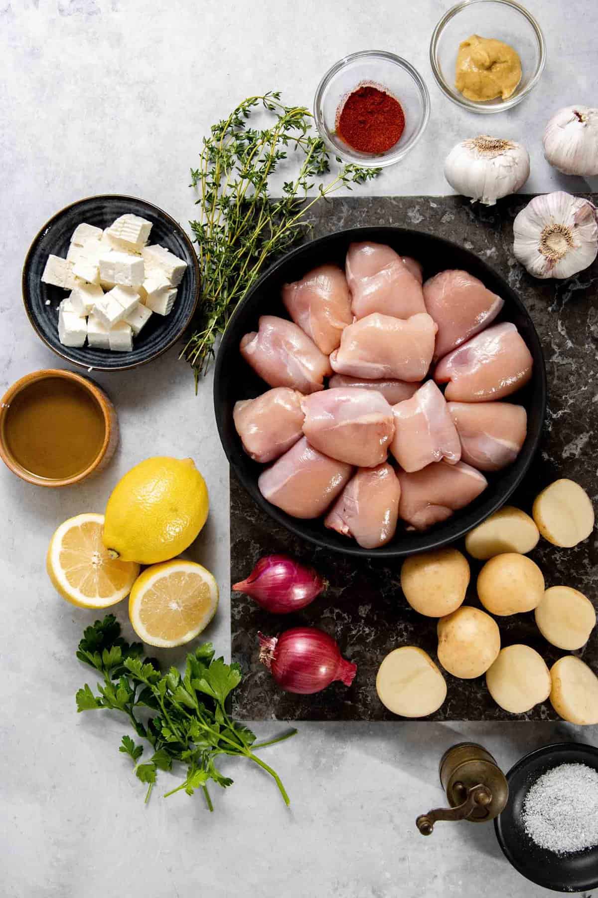 Ingredients arranged on a surface, including raw chicken pieces in a bowl, garlic, mustard, paprika, feta cheese, lemon, shallots, small potatoes, fresh herbs, and olive oil.